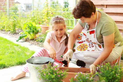 A woman and a young girl are working together in a garden outdoors, with the woman wearing a white apron with floral patterns and gardening gloves, tending to potted plants placed on a wooden deck. The girl, dressed in a light pink top and pink and white striped trousers, is assisting by handling a small gardening tool. Behind them, there is a lush green lawn, a variety of flowering plants, and a garden bed with soil and foliage. In the background, a glass greenhouse or garden conservatory is visible, surrounded by mature trees and shrubs, indicating a well-maintained outdoor space in Ruislip. The scene suggests an activity focused on gardening and outdoor plant care, characteristic of landscaping and gardening services provided by specialists like Landscapers Ruislip, with natural lighting and a peaceful atmosphere typical of a typical UK residential garden in late spring or summer.