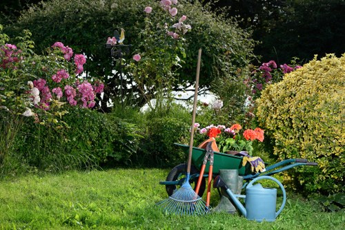 The image depicts a well-maintained garden area with a lush green lawn in the foreground, dotted with various gardening tools and equipment including a blue watering can, a rake, and a wheelbarrow filled with additional tools. Behind the tools, there are vibrant flowering bushes with pink and purple blooms, along with neatly trimmed shrubs and a small tree, creating a colourful and inviting outdoor space. The garden is bordered by taller foliage and trees, providing a natural backdrop, and the scene suggests a well-kept garden area typical of residential outdoor spaces in Ruislip. The lighting indicates a bright day, with natural sunlight illuminating the plants and equipment, highlighting the natural textures and tones of the grass, leaves, and flowers. This setting exemplifies the type of environment where professional gardening and landscaping services from Landscapers Ruislip might be used to maintain or improve outdoor areas, including lawn care and flower bed upkeep.