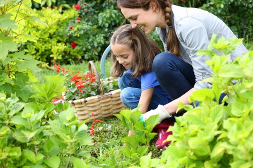 The image depicts a woman and a young girl gardening together in a lush, well-maintained backyard garden. They are both crouched down near a garden bed filled with green foliage, with the girl holding a small wicker basket, possibly for collecting flowers or garden produce. The woman is smiling and guiding the girl in the activity, which appears to involve planting or harvesting. Surrounding them are various garden elements including dense leafy shrubs, vibrant red flowering plants, and a neatly trimmed lawn with dense, green grass. In the background, there are more plants and possibly a hedge or boundary plants that frame the garden space. The lighting suggests a bright, natural outdoor setting with soft sunlight illuminating the scene, indicating fair weather. The environment emphasizes a healthy, lively outdoor space suitable for family gardening activities, reflecting high standards of outdoor maintenance and landscaping typical of professional garden care services such as those offered by Landscapers Ruislip, with subtle hints of the local garden environment in Ruislip, Middlesex.