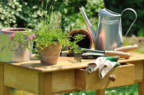A wooden outdoor garden table situated in a lush, green garden area features various gardening tools and plant-related items. On the table, there are multiple terracotta and ceramic pots filled with green herbs and flowering plants, alongside a gardening fork and trowel, both with wooden handles. A metallic watering can with a long spout is positioned at the back of the table, ready for watering plants. A pair of beige gardening gloves rests on the edge of the table, indicating recent or ongoing garden activities. The background reveals a vibrant garden setting with dense foliage, suggesting a well-maintained outdoor space in Ruislip or nearby areas. The scene captures a typical gardening moment, with natural daylight illuminating the tools and plants, supporting services such as lawn care, planting, and garden maintenance by Landscapers Ruislip, focused on enhancing outdoor spaces throughout the local postcode area.