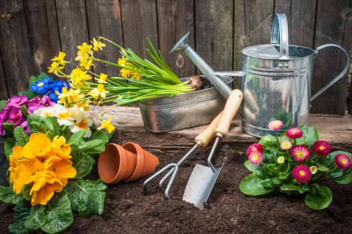 A garden scene featuring a section of soil prepared for planting with various gardening tools including a hand fork, trowel, and watering can. Brightly coloured flowering plants such as primroses and daffodils are arranged in the soil area, alongside empty terracotta flower pots. In the background, there is a weathered wooden fence that encloses the garden space. The environment suggests early spring or late winter gardening activity, with vibrant flower blooms adding natural colour contrast to the earthy soil and garden tools. The overall setting exemplifies a typical outdoor garden area suitable for planting and landscape enhancement, aligning with services offered by companies like Landscapers Ruislip in the London area, including Ruislip and surrounding postcodes. The scene emphasizes a well-maintained outdoor space ready for further landscaping or plant care, reflecting professional gardening and landscaping practices.