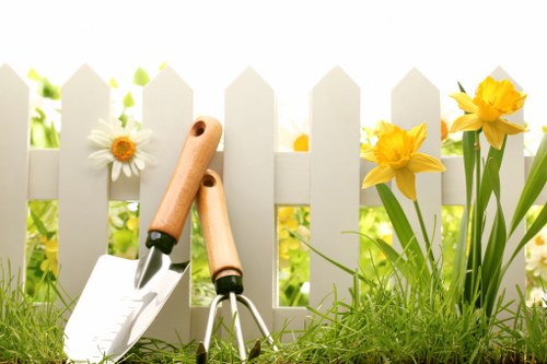 A close-up view of a tidy garden with a white picket fence in the background, featuring bright yellow daffodils and white daisies growing along the edge of a well-maintained lawn. In the foreground, there are gardening tools including a hand trowel and a small hand rake with wooden handles, resting on the grass. The grass is lush and green, with a slightly uneven surface, and the sunlight creates a warm, inviting atmosphere typical of spring or early summer. The scene suggests ongoing gardening or lawn care activity in a residential outdoor space within Ruislip, highlighting the area's typical suburban garden features. This setting underscores the importance of professional gardening services offered by companies like Landscapers Ruislip, especially for maintaining vibrant flower beds and healthy lawns in local gardens.