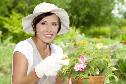 The image depicts a woman in a garden setting with vibrant greenery and colorful flowers. She is smiling and wearing a wide-brimmed white hat, a white shirt, and gardening gloves, suggesting she is engaged in gardening activities. In her hands, she holds a small terracotta pot filled with pink and white flowering plants, possibly geraniums, which are positioned centrally in the scene. The background shows a landscaped outdoor space with well-maintained grass, shrubs, and trees, indicating a lush garden environment typical of residential outdoor spaces in Ruislip or nearby areas. The weather appears to be bright and sunny, with natural light illuminating the scene, highlighting the health of the plants and the woman's cheerful demeanor. This image captures a moment of outdoor gardening, aligning with professional gardening services offered by Landscapers Ruislip, especially around sustainable planting and garden care for residential properties in the local area.