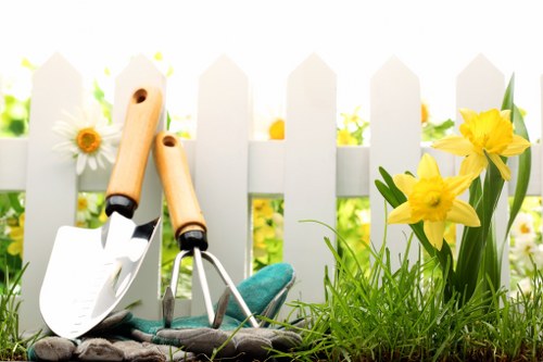 The image depicts a small garden scene with a white wooden picket fence in the background, behind which a lush outdoor space is visible. In the foreground, there are two small gardening tools placed on the soil: a trowel with a wooden handle and a hand rake, both with metallic heads, resting among healthy, green grass and young yellow daffodil flowers in bloom. To the left of the tools, a white daisy with a yellow center is partially visible, also growing near the fence. The garden area appears well-maintained, showcasing a combination of neatly edged flower beds and a vibrant, green lawn. The scene suggests a mild, sunny day ideal for outdoor gardening activities, highlighting elements typical of a residential garden in Ruislip or nearby locations within the HA postcode district. The professional landscaping company, Landscapers Ruislip, offers gardening and outdoor maintenance services to keep such garden spaces attractive and healthy, aligning with local landscaping standards.