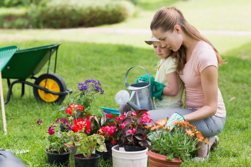 A woman and a young girl are kneeling on a well-maintained, lush green lawn in a backyard or garden area, engaged in gardening activities. The woman is assisting the girl with watering plants using a small watering can, both focused on a collection of vibrant flowering plants in various pots, including red, purple, and orange blossoms. In the background, there are trees and a pathway, suggesting an outdoor space designed for leisure and gardening. To the left, a green garden wheelbarrow with yellow wheels is visible on the grass, indicating ongoing gardening or lawn care tasks. The scene is brightly lit, likely in natural daylight, with a soft, sunny atmosphere suitable for outdoor gardening work, reflecting professional landscaping care typical of services provided by Landscapers Ruislip, serving local communities in Ruislip or nearby areas. This image captures the peaceful, productive environment of a well-maintained garden ready for routine upkeep or renovation undertaken by gardening professionals.