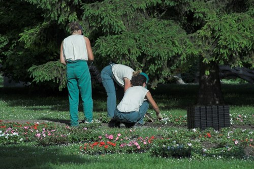 The image depicts three individuals working together in a lush, green outdoor garden space, likely part of a landscaped yard, possibly in Ruislip or nearby. Two of them are crouching and engaged in planting or tending to a flower bed that features a variety of small, vibrant pink, red, and white flowers arranged along the edge of a well-maintained grassy lawn. The third person stands nearby, observing or assisting. The garden is bordered by mature trees with dense green foliage, providing shade and a natural backdrop. The ground appears moist and fertile, with dark soil visible in areas where planting occurs. In the background, dense tree branches and leaves create a canopy that filters sunlight, casting gentle natural light over the scene. The setting suggests outdoor gardening activities typical of residential landscaping services offered by companies like Landscapers Ruislip, emphasizing garden maintenance, planting, and outdoor beautification, all within a peaceful and inviting garden environment.
