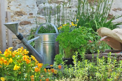 The image depicts a small, neatly maintained garden area with a variety of plants and flowers. In the foreground, there is a cluster of vibrant yellow and orange flowering plants, likely annual or perennial species, adding bright colour to the scene. Behind these flowers, a metal watering can with a curved handle is prominently placed, suggesting active gardening or horticultural care. To the right, a lush green potted plant with broad, healthy leaves is visible, alongside other smaller plants that are neatly arranged in containers. The background features a textured stone wall, typical of outdoor garden boundaries in Ruislip, with tall, grassy plants and shrubs growing along the wall, providing natural height variation and privacy. A garden tool may be partially visible, and the overall scene appears to be set during a sunny day with natural lighting illuminating the garden, reflecting the characteristic outdoor aesthetic managed by services like those from Landscapers Ruislip. The composition showcases a well-tended, engaging outdoor space combining both cultivated plants and decorative elements to support gardening and landscaping efforts in the local area.