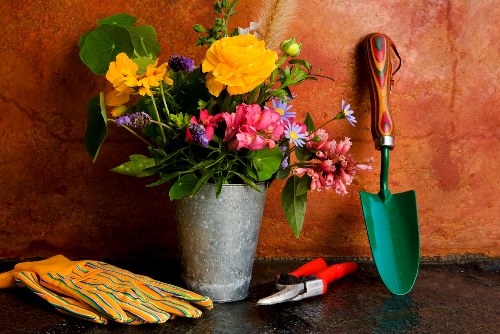 A close-up view of a gardening workspace featuring a large grey concrete pot filled with a colorful arrangement of fresh flowers, including yellow, pink, purple, and blue blooms, placed on a dark outdoor surface against a textured reddish-brown wall. To the right of the pot, there is a small green garden trowel with a wooden handle, resting upright on the ground. Adjacent to the trowel, a pair of red-handled pruning shears is lying closed on the surface. To the left of the pot, a pair of yellow and blue striped gardening gloves is folded neatly. The scene appears to be set in an outdoor garden area, reflecting a routine gardening activity with tools and plants ready for outdoor maintenance, aligning with the gardening services offered by Landscapers Ruislip in the local area of Ruislip, Middlesex. The natural lighting suggests a sunny day, highlighting the vibrant colors of the flowers and the gardening tools.