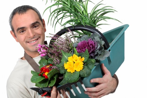 A professional gardener from Landscapers Ruislip, dressed in a light-colored shirt, is smiling and holding a small, plastic garden basket filled with a variety of colorful flowering plants, including purple, yellow, red, and pink blooms with green foliage. The background features a lush, well-maintained garden with a dense hedge, a wooden garden trellis, and a garden shed painted in a greenish shade. The scene is outdoors during daylight, with natural light illuminating the plants and the gardener, emphasizing the healthy condition of the garden plants and the neat organization of the outdoor space, which is typical of gardening and landscaping services in the Ruislip area.