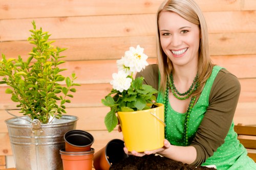 The image shows a young woman with blonde hair and a bright smile sitting outdoors in a garden setting. She is wearing a green polka-dot apron over a brown long-sleeved top, and accessorized with green beaded necklaces, indicating engagement in gardening activities. In front of her on the table are several potted plants, including a small shrub with vibrant green leaves in a metal bucket, and a flowering plant with white blossoms in a yellow pot, along with empty plant pots nearby. The background features a wooden fence, suggesting a private backyard or garden area typical of gardens in Ruislip. The scene is well-lit, likely during a clear day, emphasizing the natural tones of the plants, pots, and wooden backdrop. The woman’s focused and cheerful expression reflects enjoyment of gardening work, which aligns with professional landscaping and gardening services offered by Landscapers Ruislip. The image conveys a professional yet casual outdoor gardening environment, suitable for illustrating garden care, planting, and outdoor maintenance services in local gardens or landscaped yards.