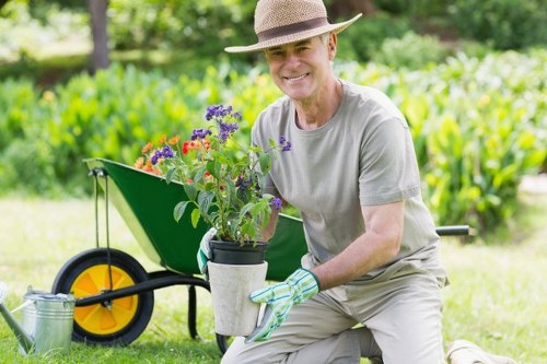 A smiling adult man wearing a straw hat, grey t-shirt, and gardening gloves kneels on a well-maintained lawn in a garden, holding a container with purple and red flowering plants. Behind him, a black and yellow wheelbarrow is positioned on the grass, partially filled with gardening tools and additional plants, with a clear, bright outdoor environment featuring lush green foliage and trees in the background. The garden area includes a neatly edged flower bed with shrubs, and the scene is illuminated by natural daylight, suggesting good weather. The image highlights outdoor garden maintenance and planting activities, aligning with gardening services offered by companies such as Landscapers Ruislip in the area around Ruislip, Middlesex, which emphasizes professional lawn care, planting, and garden landscaping near local Hillingdon postcode zones.