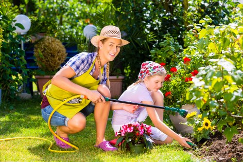 In a lush residential garden with a well-maintained lawn and vibrant flower beds, a woman and a young girl are engaged in gardening activities. The woman, wearing a straw hat, a checked shirt, and a bright yellow apron, is crouched down holding a garden hose, smiling as she supervises the girl. The girl, dressed in a light top and pink rain boots, is focused on planting or tending to the colorful flowers and plants near a flower bed with red, yellow, and pink blooms. The garden features a hedge and shrubbery in the background, with decorative garden lanterns and lush greenery creating a welcoming outdoor space. The scene appears to be sunny, with natural light illuminating the garden, highlighting the natural tones of the soil, grass, and vibrant plants. This outdoor environment showcases a well-cared-for garden that could benefit from professional gardening and landscaping services, such as lawn care, planting, and garden maintenance, offered by specialists like Landscapers Ruislip, enhancing the visual appeal and functionality of suburban gardens in the Ruislip area, inferred from the UK postcode HA4.