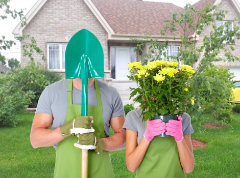 A man and woman standing in a residential garden during daytime, both wearing green aprons and gardening gloves, with the man holding a bright turquoise garden spade with a rounded blade, and the woman holding a large pink pot of yellow flowering plants, possibly chrysanthemums or similar, with green foliage. In the background, there is a well-maintained lawn, edged by a variety of shrubs and small trees, and a row of suburban houses with brick exteriors, white-framed windows, and sloped roofs. The garden area appears to be neatly landscaped with a clear lawn and organized planting beds, suggesting ongoing gardening or landscaping work typical in Ruislip or nearby areas. The overall scene highlights outdoor garden maintenance and planting activities, emphasizing the natural colors of the grass, plants, and flowers, set under natural daylight with a partly cloudy sky.