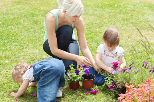 A woman with short blonde hair, wearing a light blue sleeveless top and dark blue jeans, is kneeling on a well-maintained, lush green lawn in a garden. She is engaged in planting or tending to potted plants that include purple, pink, and green foliage. To her side, two young children are actively involved; one is bending forward, reaching towards the soil with her hands, while the other, a girl with short brown hair, is sitting on the grass holding a small pink flower. The garden features a neatly bordered flower bed with a variety of colorful blooms, suggesting a vibrant, cultivated outdoor space. The environment appears bright and sunny, indicating clear weather, with natural light illuminating the scene. The background hints at a private residential garden, reflecting typical gardening and outdoor maintenance activities. This scene exemplifies family-friendly gardening and landscaping work that local Ruislip gardening services, such as those offered by landscapersruislip.co.uk, might support.