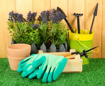 The image showcases a neatly arranged garden scene with gardening tools and plants. In the foreground, there are several purple flowering lavender plants in small, black plastic pots positioned behind a wooden garden box. To the left, there are two empty terracotta pots, and next to them, a pair of bright turquoise gardening gloves rests on a wooden platform with small garden tools. In the background, a bright yellow plastic bucket contains longer-handled garden tools, including a spade, rake, and trowel, all standing upright. The garden setting features a vibrant green artificial grass surface, with a wooden fence or paneling in the background, suggesting a well-maintained backyard space typical of Ruislip. This scene is indicative of outdoor maintenance and garden preparation activities, aligning with professional services provided by Landscapers Ruislip for comprehensive gardening and landscaping solutions, including plant care, lawn edging, and outdoor equipment organization.